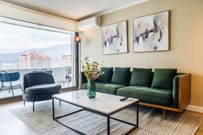 A modern living room with a green sofa, a coffee table, and a vase with flowers. There are two abstract paintings on the wall and a city view through the window. - Luxury 2 Bedroom Apartment with Pool and Parking (Las Condes)