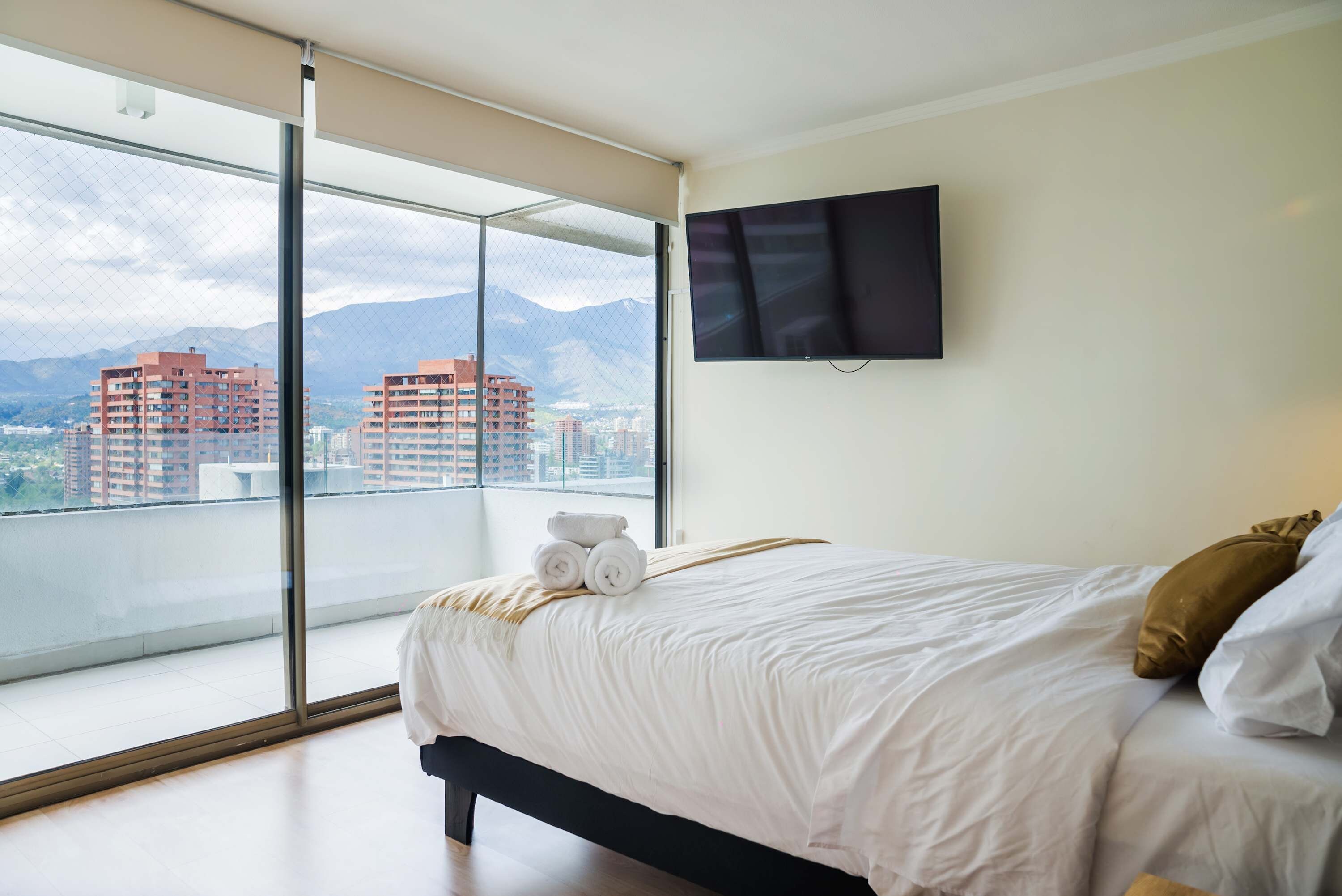 A hotel room with a large window, a bed, a flat-screen TV, and a view of buildings and mountains.