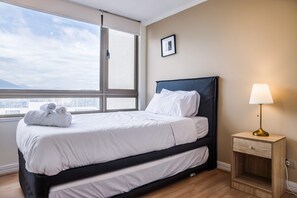 A hotel room with a bed, a nightstand, a lamp, and a window with a view of the city. - Luxury 2 Bedroom Apartment with Pool and Parking (Las Condes)