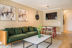 A living room with a green sofa, a coffee table, abstract paintings, a TV, and a dining area with red chairs. - Las Condes Modern Apartment 2BR2BA, Pool & Views (Las Condes)
