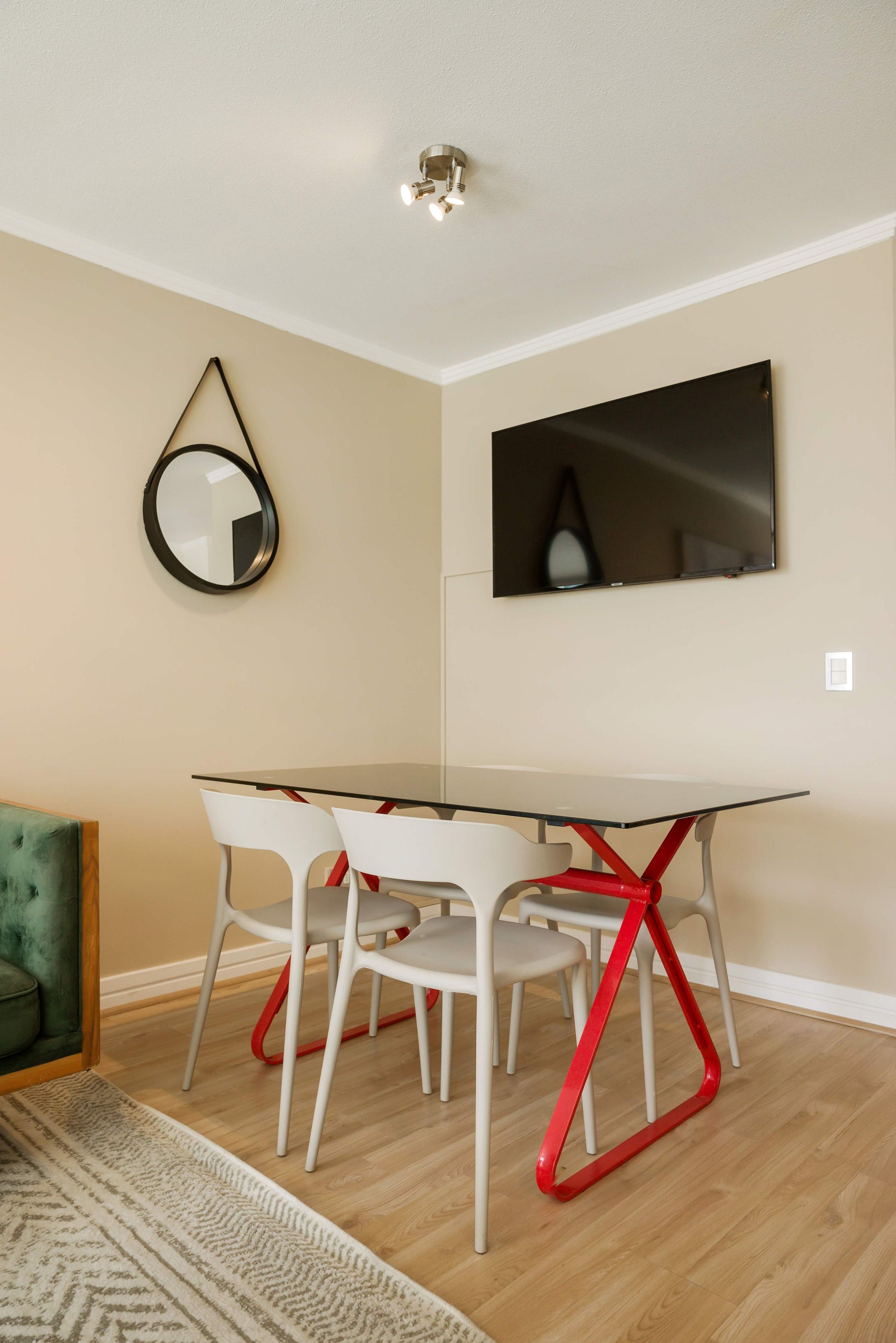 A dining area with a glass table, white chairs, a red chair frame, a round mirror, and a flat-screen TV on the wall.