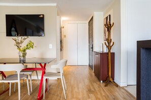 A modern living room with a flat-screen TV mounted on the wall, a dining table with chairs, a vase with flowers, and a coat rack. - Luxury 2 Bedroom Apartment with Pool and Parking (Las Condes)