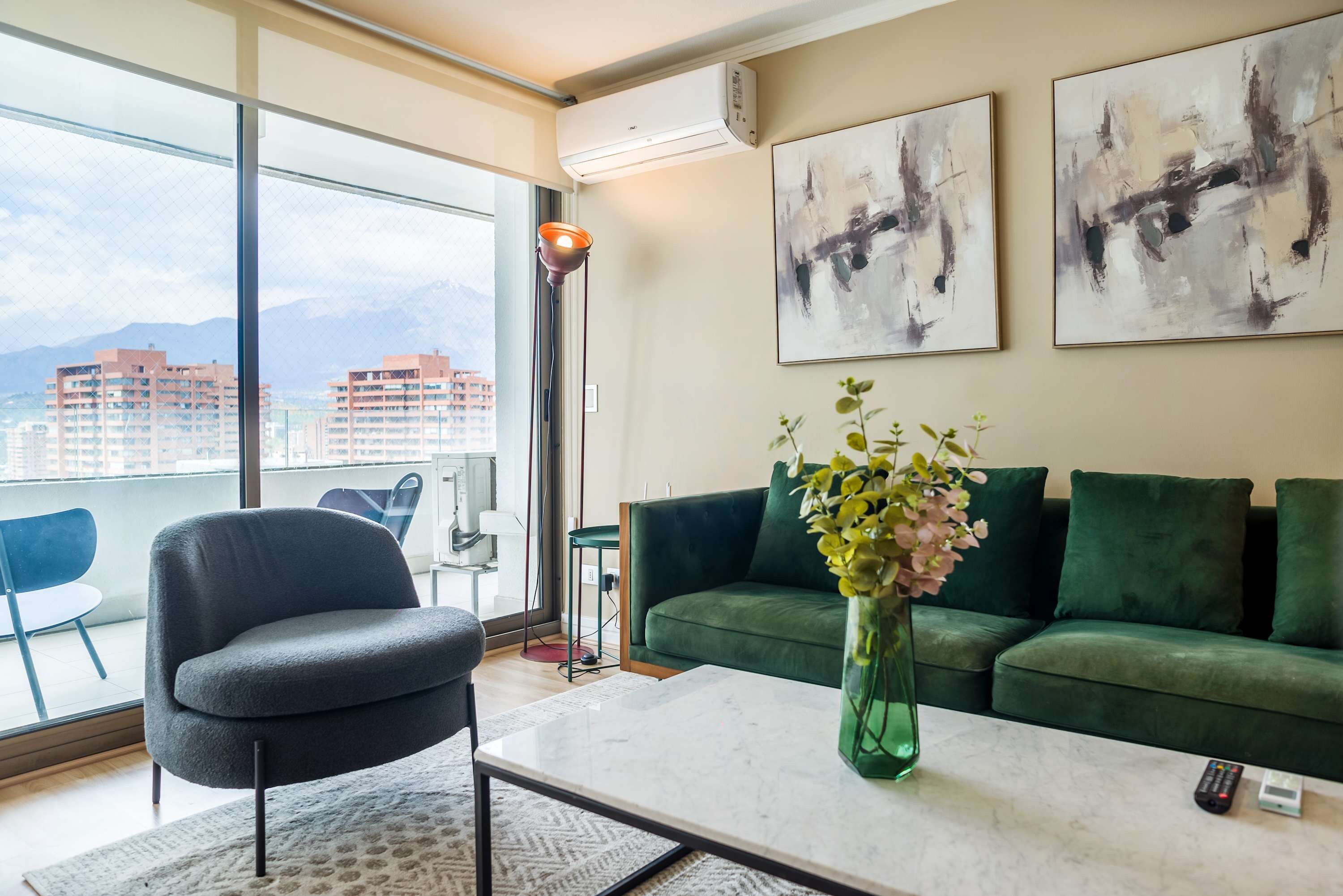 Ein modernes Wohnzimmer mit einem grünen Sofa, einem runden Sessel, einem Couchtisch und einer Vase mit Blumen. An den Wänden sind abstrakte Gemälde und es gibt ein großes Fenster mit Blick auf Gebäude.