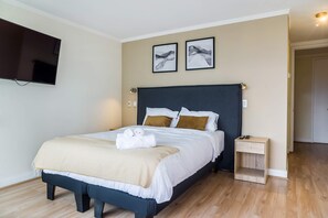 Una cama bien tendida con sábanas blancas y un plaid color beige, una mesita de noche con lámpara, y dos cuadros enmarcados en la pared.