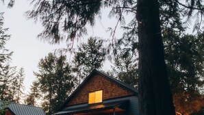 Una cabaña con techo inclinado, un porche y un gran árbol en primer plano.