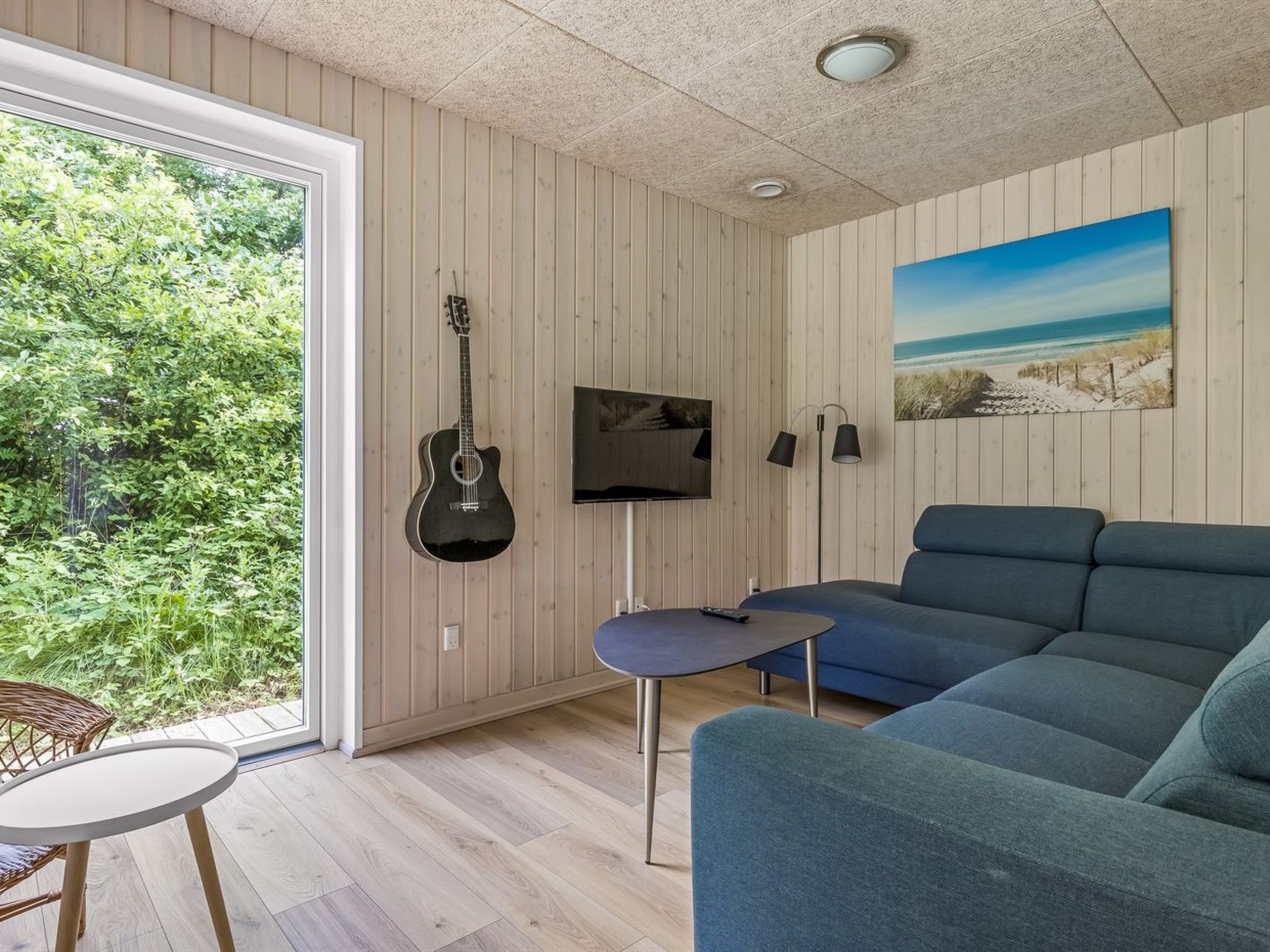 A modern living room with a large window, a blue sofa, a small table, a guitar, a TV, and a painting.