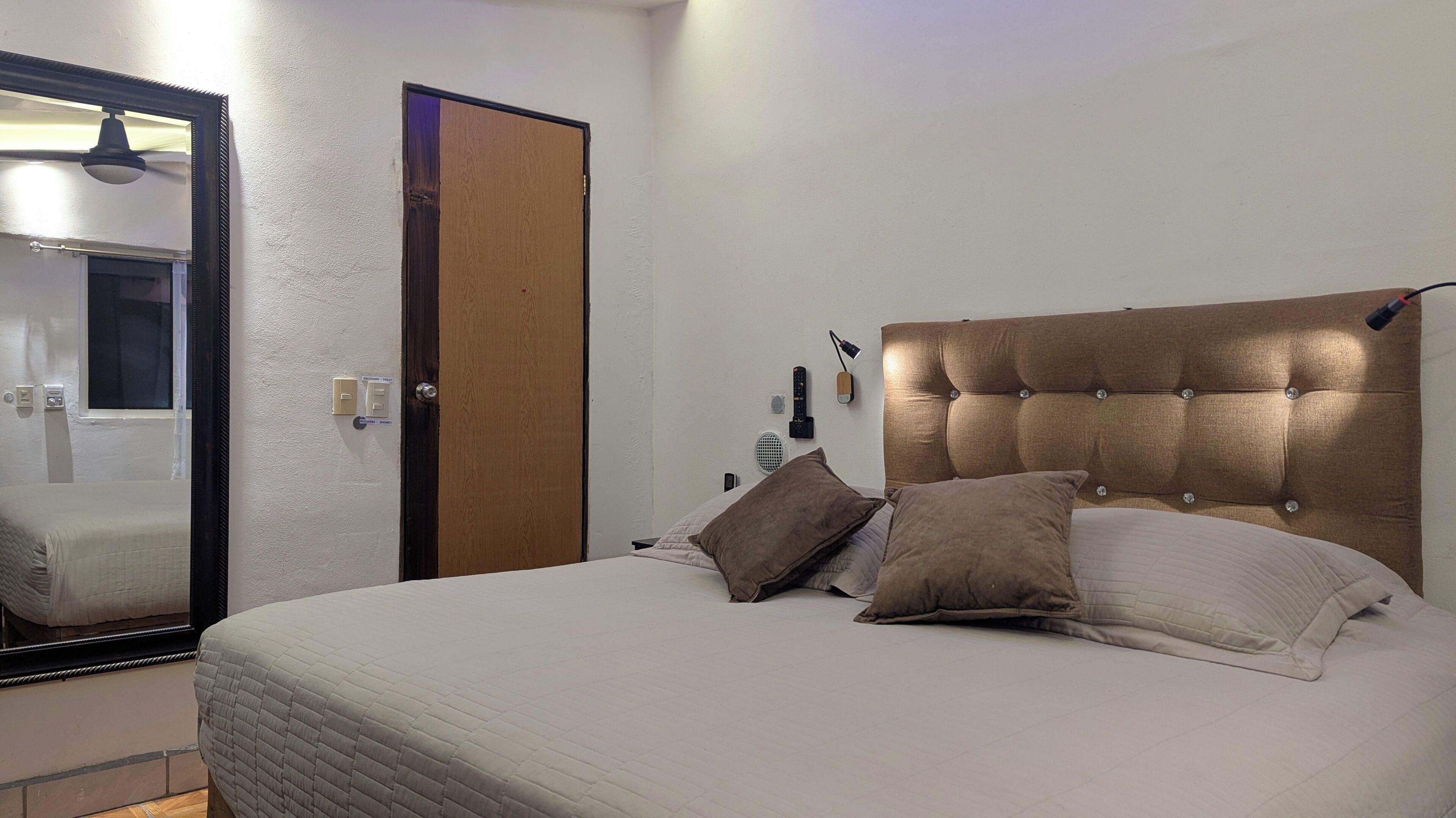 A neatly made bed with a tufted headboard, a large mirror, and a door with a wooden panel.
