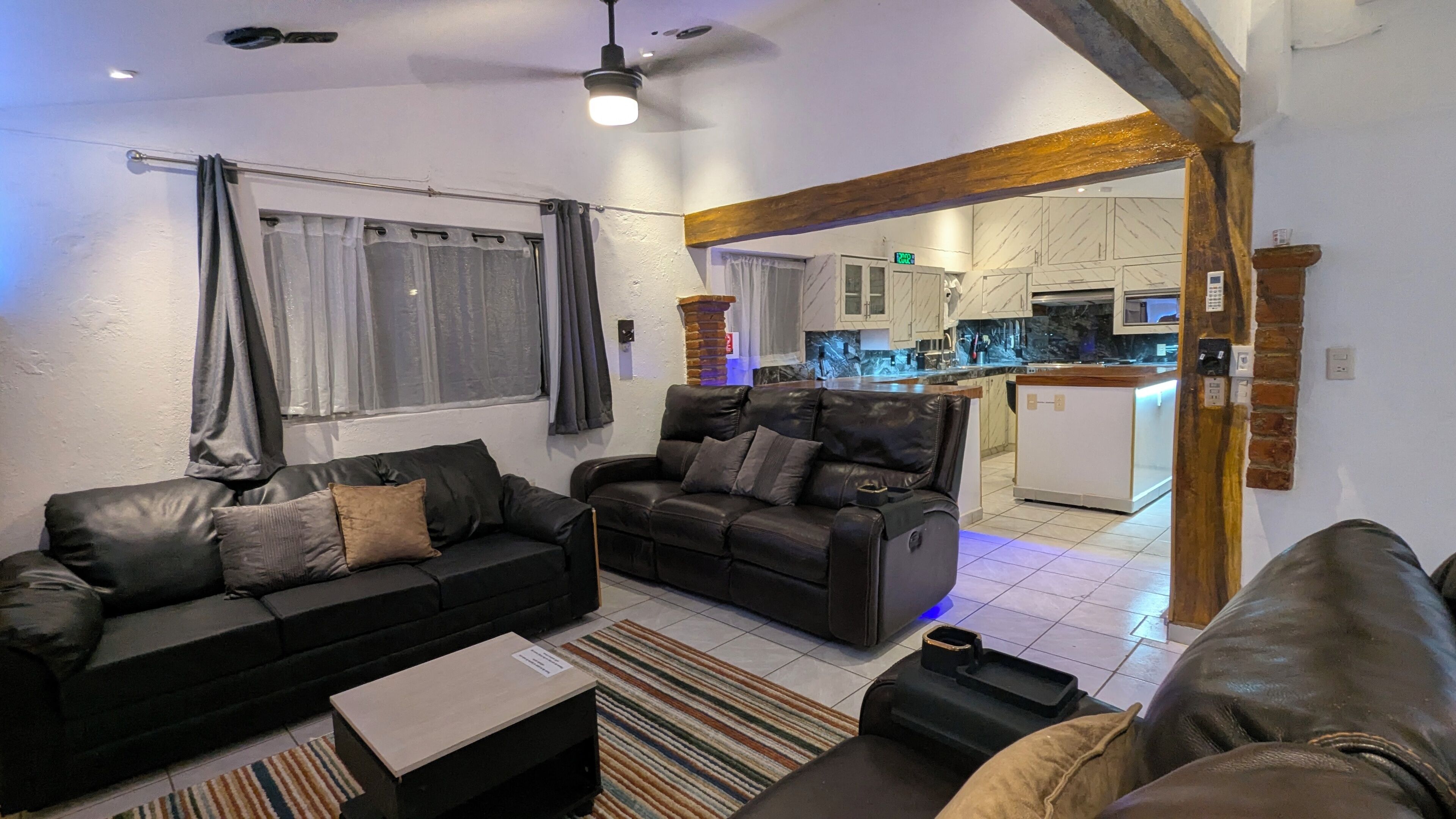 A living room with a sofa, a coffee table, and a rug. There is a kitchen visible in the background.