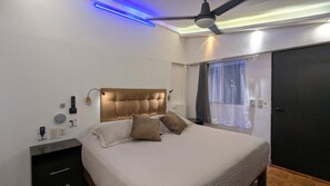 Quarto com cabeceira estofada, cama com almofadas, mesa de cabeceira com abajur, ventilador de teto e janela com cortinas.