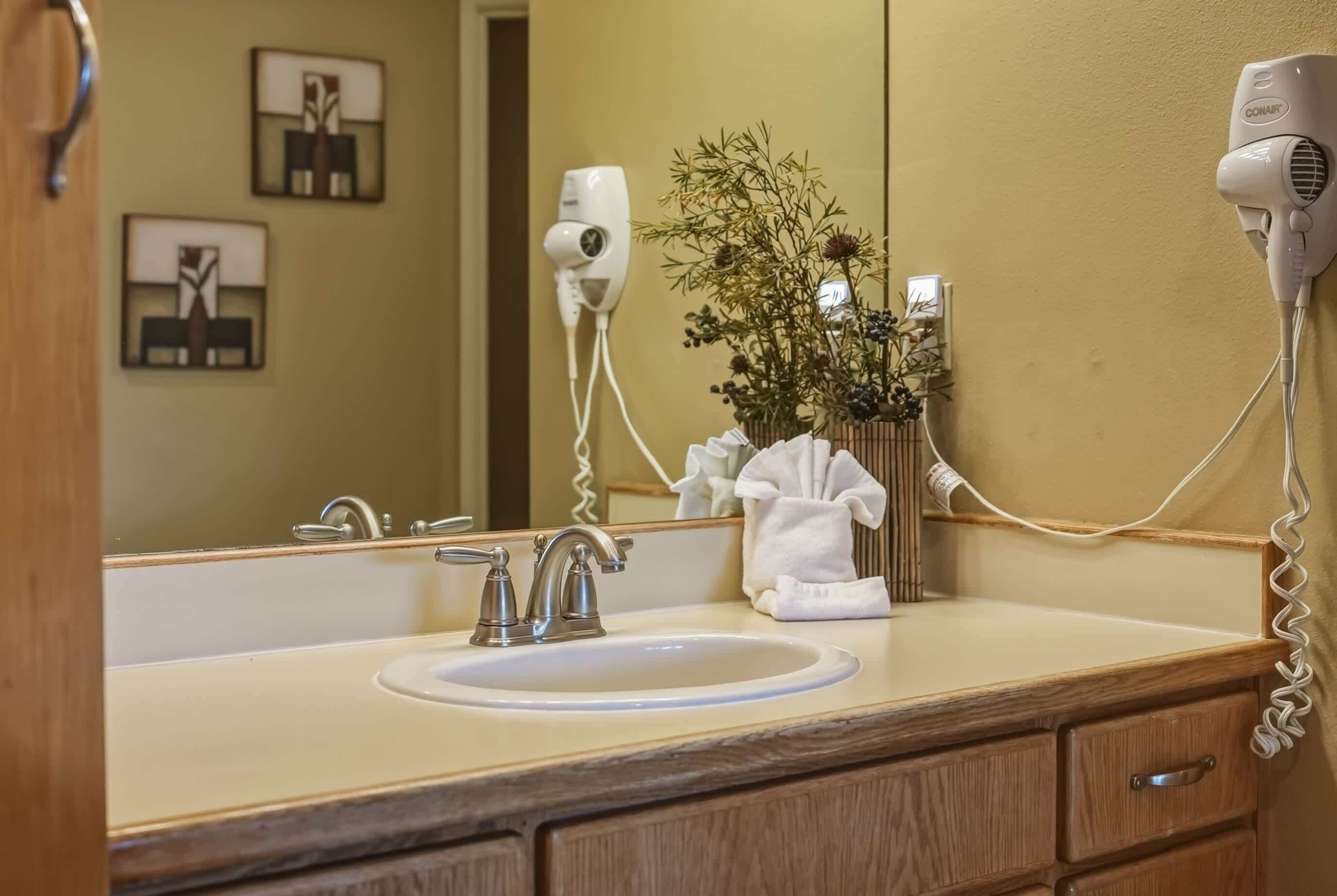 Une salle de bain équipée d’un lavabo, d’un miroir, d’un sèche-cheveux et d’un vase avec des fleurs.