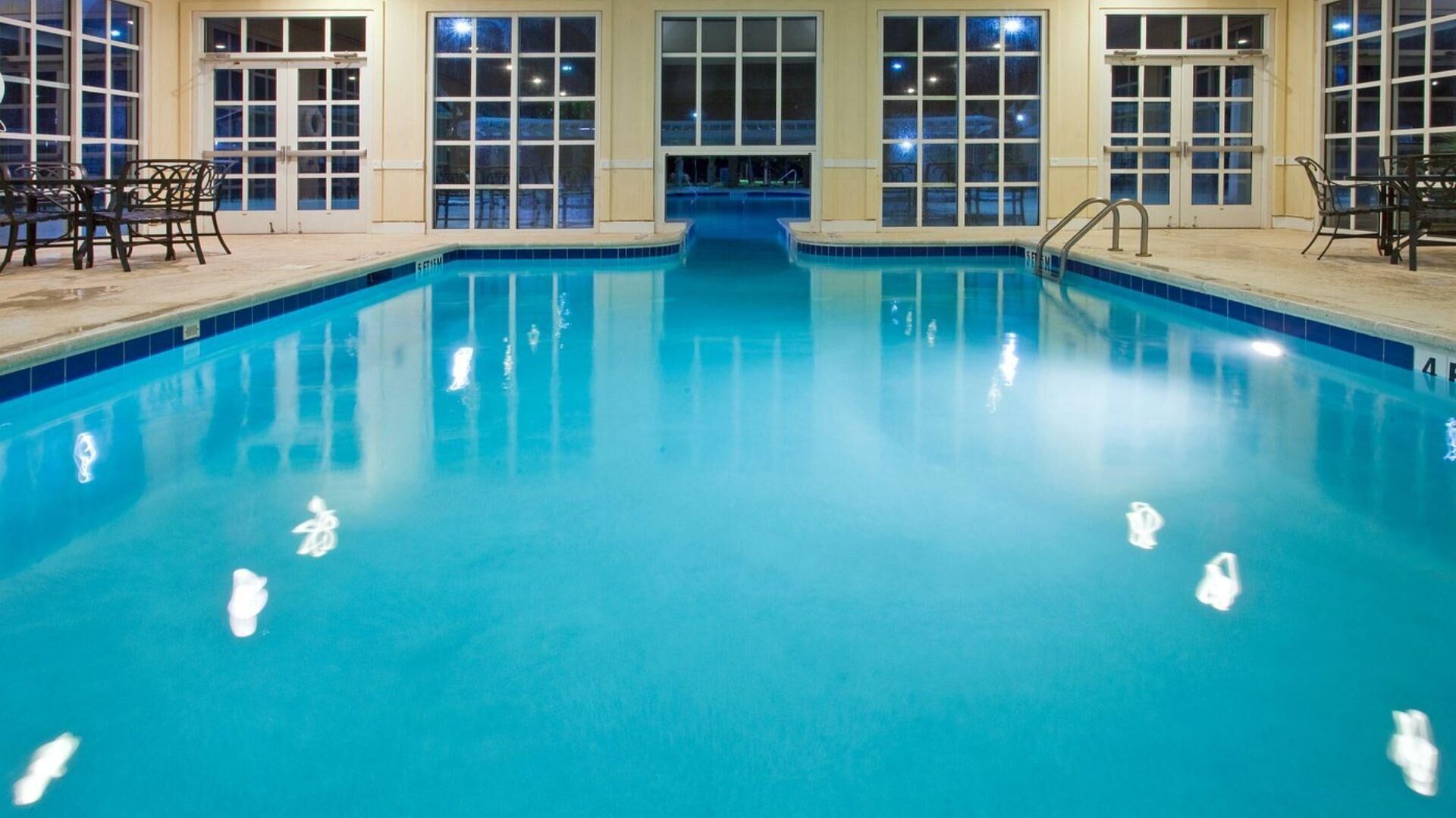 An indoor swimming pool with clear blue water, surrounded by large windows and doors, and equipped with metal handrails.