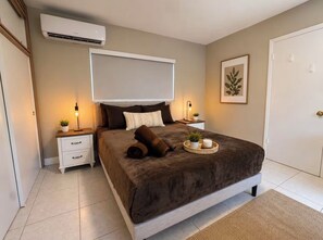 Ein Schlafzimmer mit einem Bett, Nachttischen, einer Klimaanlage, einer Tür und einem gerahmten Bild an der Wand.