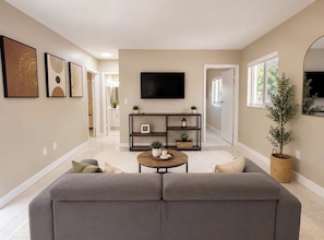 Ein Wohnzimmer mit grauem Sofa, Couchtisch, Fernseher an der Wand und einer Bücherwand mit Pflanzen.