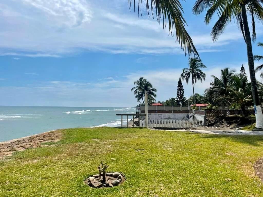 A grassy area with a small rock formation, palm trees, and a building with a red roof near the ocean.