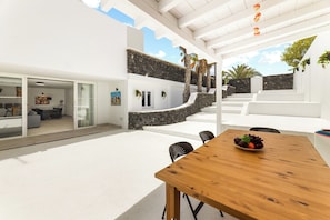 A modern outdoor dining area with a wooden table and chairs, a stone wall, and a white building with glass doors. - Home2Book Elegant Villa with Jacuzzi Costa Teguise (Teguise)