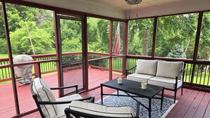 Uma área externa coberta com deck vermelho, ventilador de teto, sofá branco e mesa com duas xícaras.