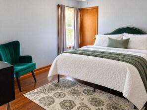 A bedroom with a bed, a chair, a window with curtains, and a patterned rug.