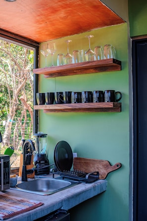 Une cuisine avec une étagère en bois où sont posés des verres à vin et des tasses, une machine à café et une planche à découper.