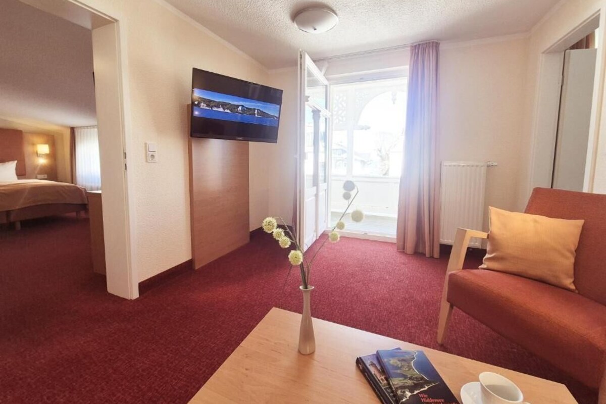 A hotel room with a bed, a sofa, a coffee table with a vase and a book, a flat-screen TV mounted on the wall, and a view through the door.