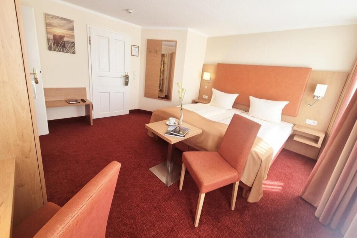 A hotel room with a bed, a desk, a chair, a small table, a mirror, and a door.