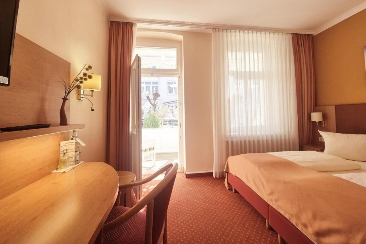 A hotel room with a bed, a desk, a chair, a lamp, and a view of the outside.