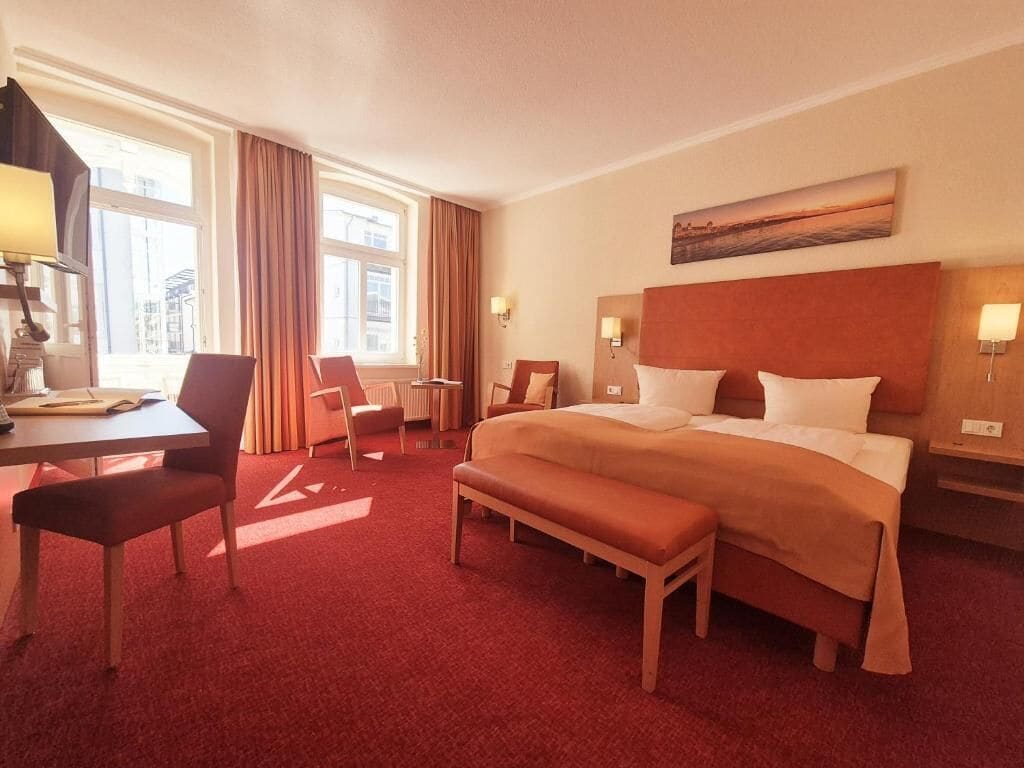 A hotel room with a large bed, a desk, a chair, a bench, and a view of the city through the window.