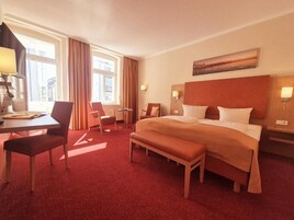 Ein Hotelzimmer mit einem großen Bett, einem Schreibtisch, einem Stuhl, einer Bank und Blick auf die Stadt durch das Fenster.
