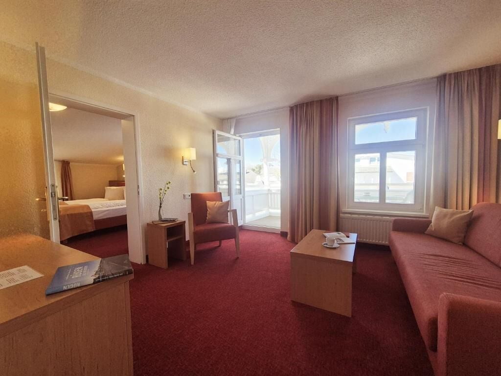 A hotel room with a bed, a sofa, a coffee table, a chair, and a desk. There is a window with curtains and a view of the outside.