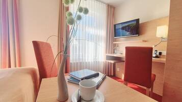 Una habitación de hotel con una mesa de madera, una taza blanca, un florero con flores verdes, un televisor, un escritorio y una silla.