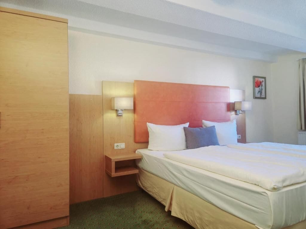 Habitación de hotel con una cama, lámparas de noche, un armario de madera y una mesita auxiliar pequeña.