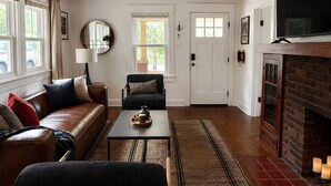 Una sala de estar con un sofá de cuero marrón, un sillón negro, una mesa de centro, una chimenea y un ventilador de techo.