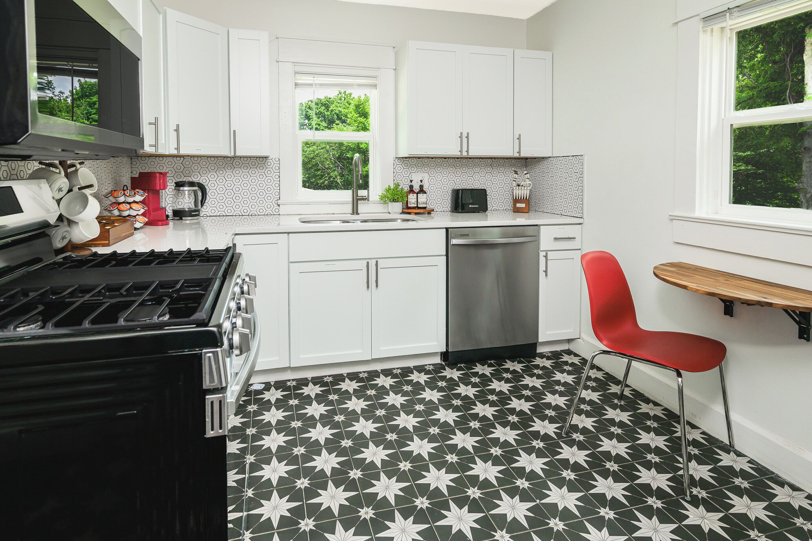 Une cuisine moderne avec des meubles blancs, un sol à motifs noir et blanc, une chaise rouge et une fenêtre donnant sur un paysage verdoyant.