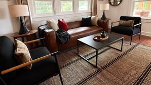 Una sala de estar con un sofá de cuero, un sillón negro, una mesa de centro y dos lámparas. Se observan ventanas con cortinas, un espejo en la pared y una alfombra en el suelo.