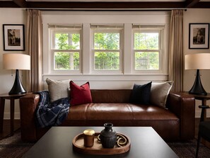 A room with a leather sofa, two side tables with lamps, a coffee table with decorative items, and a window with curtains. - Located in the IU Tailgate Fields - Across from Memorial Stadium (Bloomington)