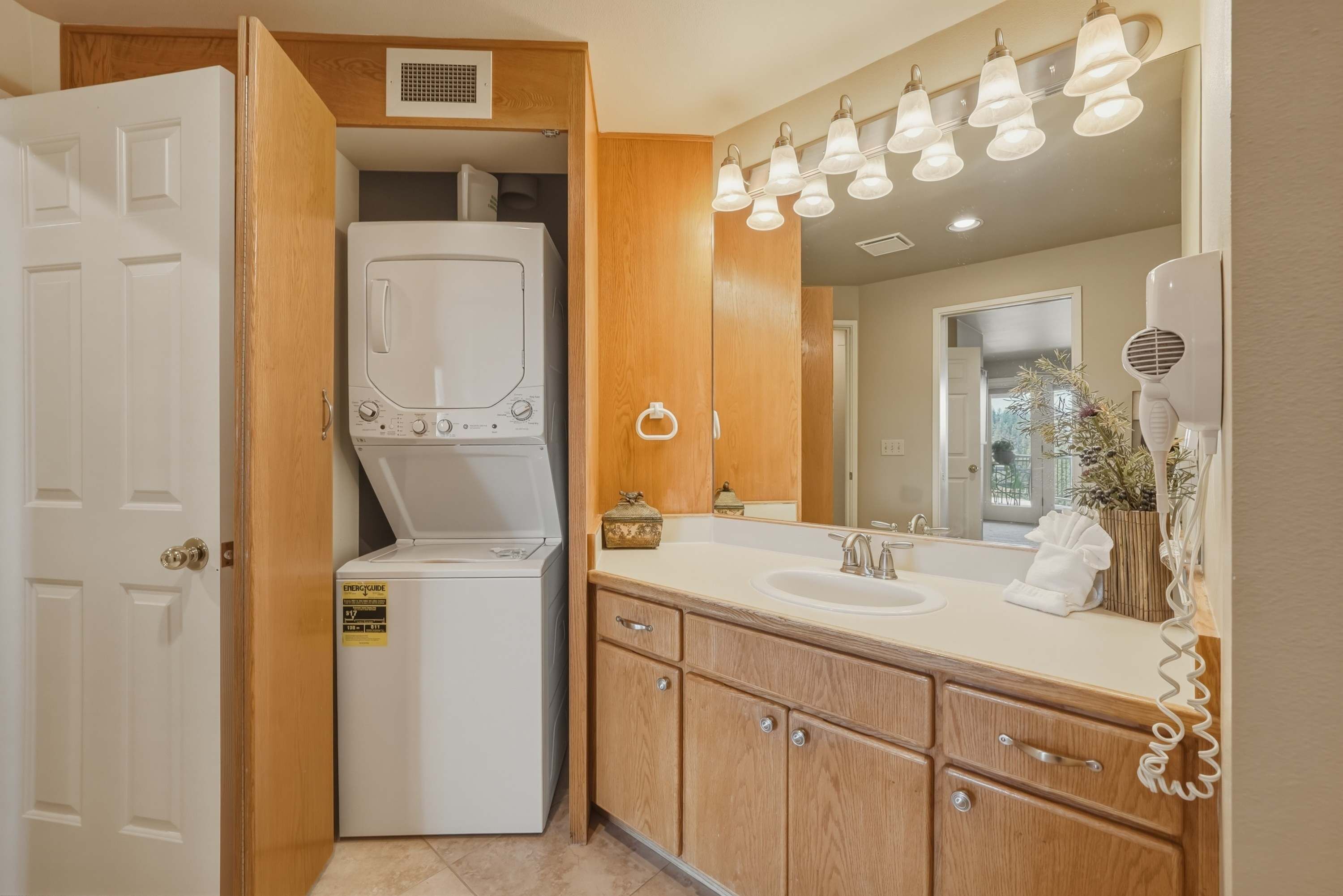A compact kitchen with a built-in washer and dryer, wooden cabinets, a sink, and a mirror with lights above.