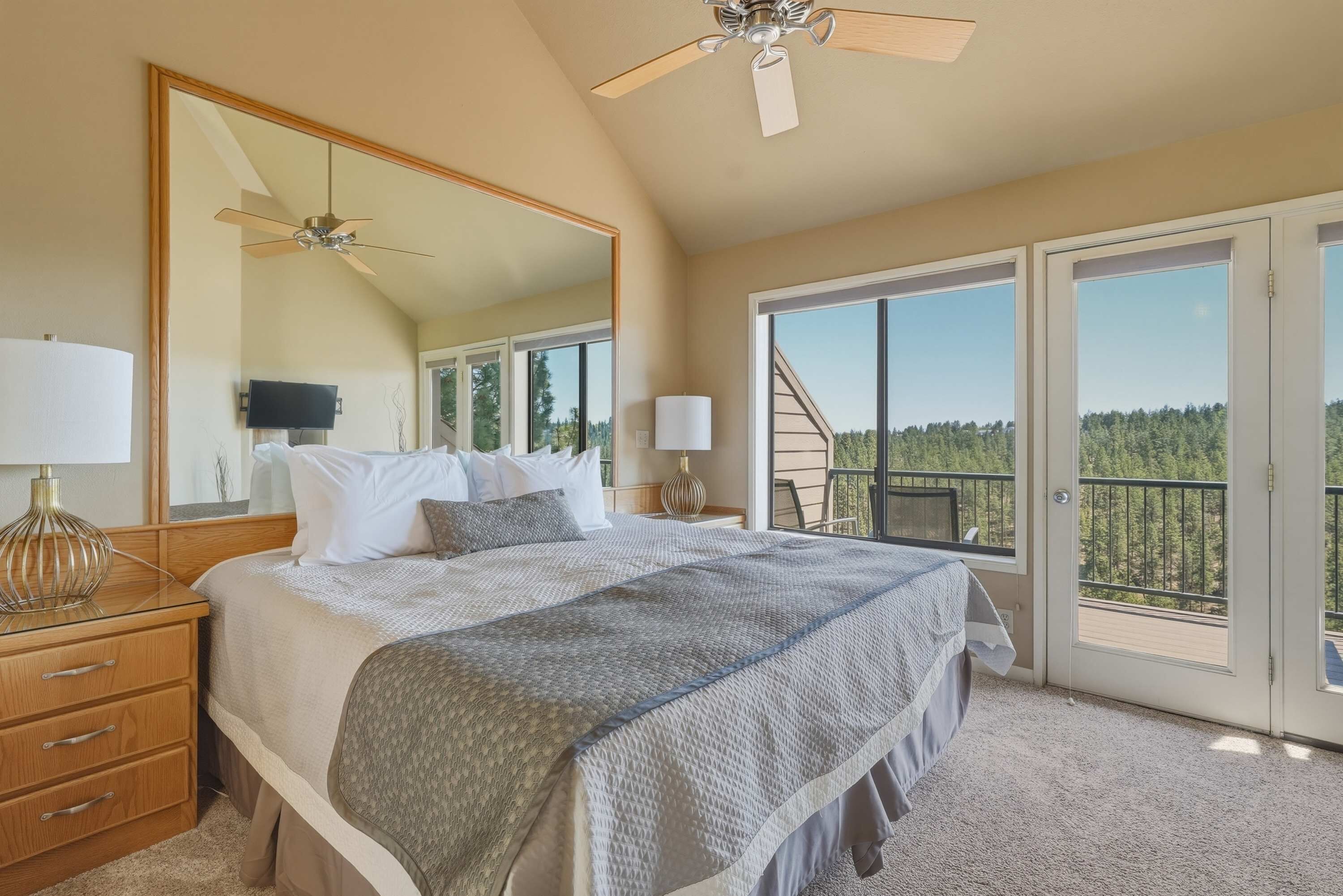 A bedroom with a large bed, a ceiling fan, a TV, and a balcony with a view of trees.