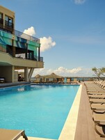Una piscina con sillones de descanso, un edificio con balcones y un cielo despejado.