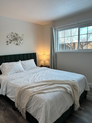 A bedroom with a bed, a wall-mounted light, a window with a view, and a decorative wall piece.