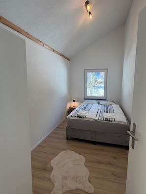 Ein schmaler Raum mit einem Bett, einem Fenster, einem Nachttisch und einem Fellteppich.