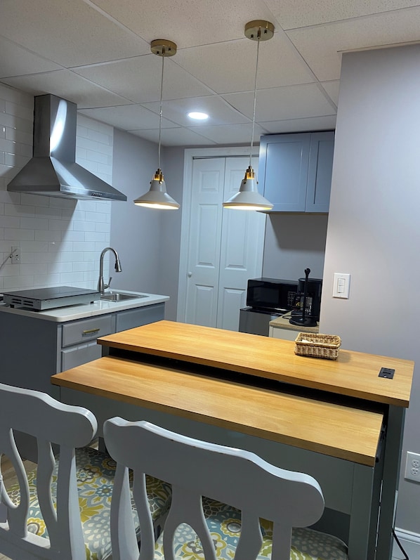 Ett kompakt kök med modern design, utrustat med en fläktkåpa i rostfritt stål, en integrerad diskho och ett matbord i trä med tillhörande stolar.