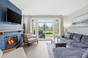 A modern living room with a fireplace, a sofa, a TV, and a view of a green landscape through large windows. - Poolside Fairway Escape | Hot Tub & Golf (The Blue Mountains)