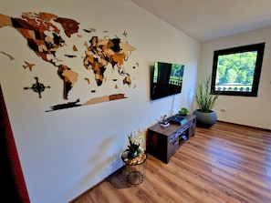 Un salon moderne agrémenté d’une décoration murale représentant une carte du monde, d’un téléviseur à écran plat, d’un parquet, d’une console sur laquelle sont disposés des objets décoratifs, et d’une grande fenêtre offrant une vue sur la verdure.