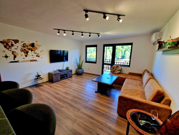 Un salon moderne avec un téléviseur à écran plat, une table basse, un canapé et une table à manger. De grandes fenêtres offrent une vue sur la verdure environnante.