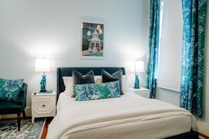 A bedroom with a bed, two bedside tables, a chair, a lamp, a painting, and curtains. - Sugar`s Inn Room #4 | Romantic Stay Near Galveston Cruise Port & Downtown (Galveston)