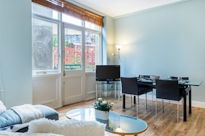 Ein Wohnzimmer mit Sofa, Couchtisch, Esstisch mit Stühlen, Fernseher und Blick auf Gebäude.