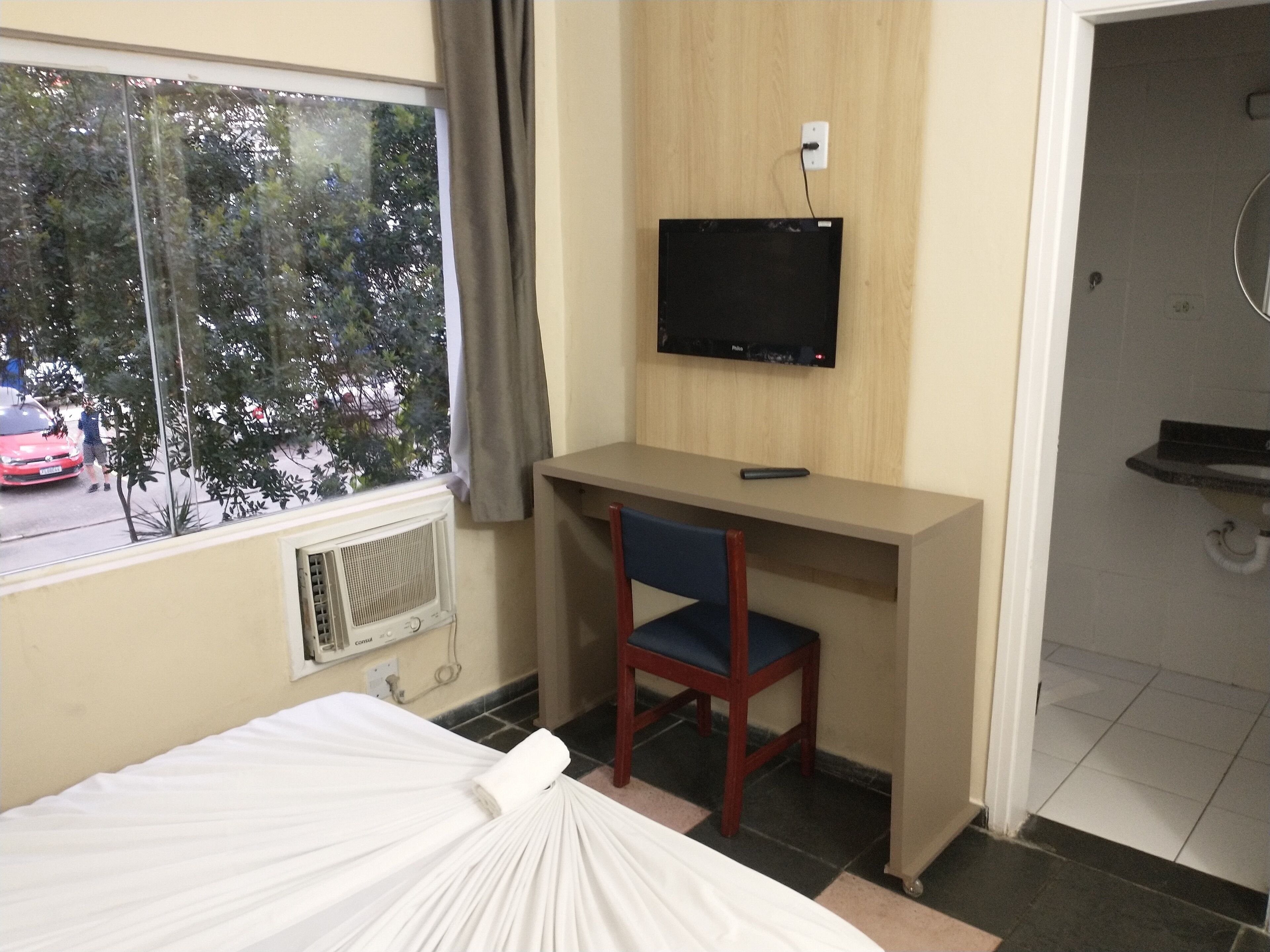 Ein Hotelzimmer mit Bett, Schreibtisch mit Stuhl, Fernseher und Fenster mit Blick auf die Straße.
