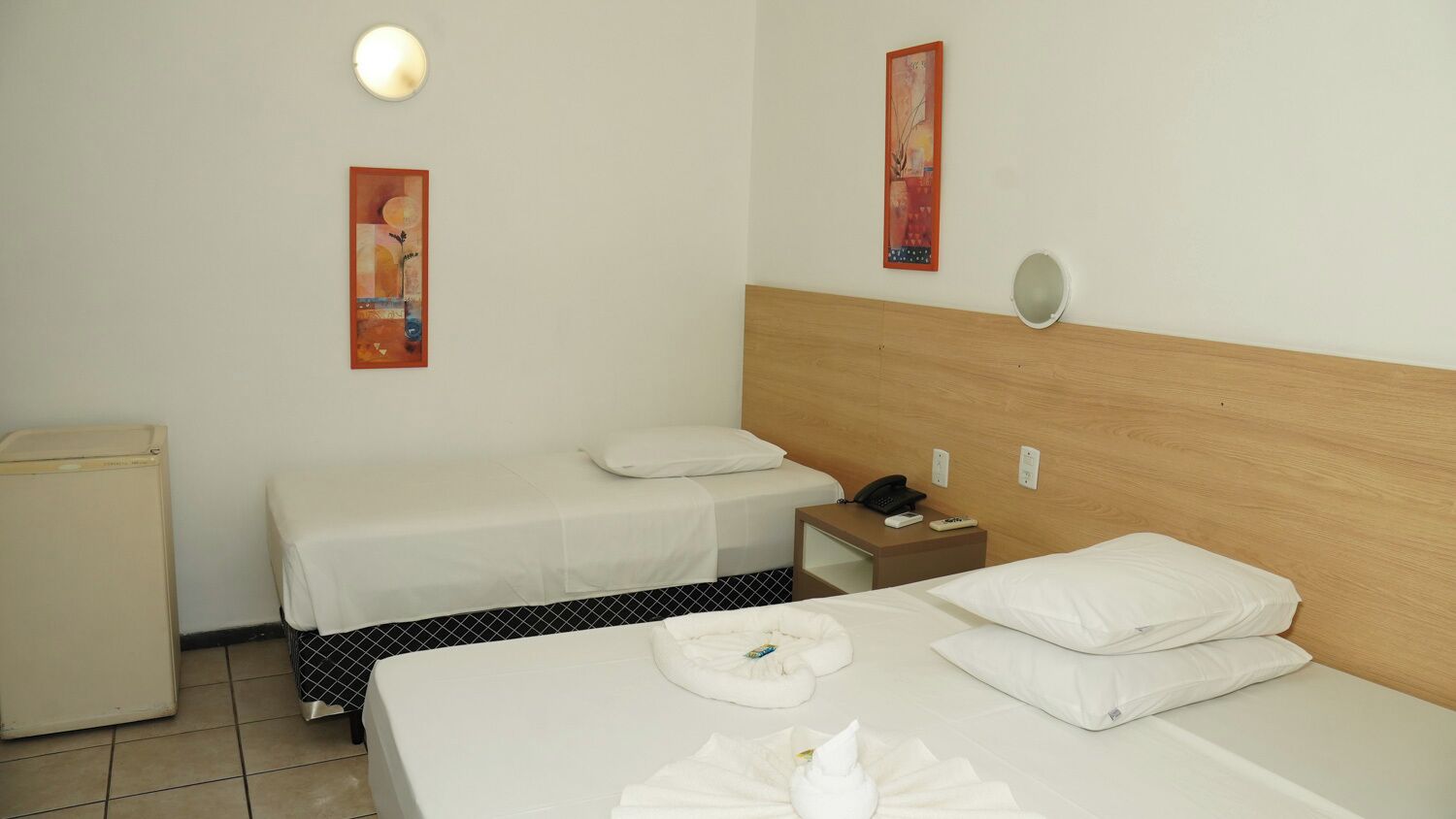 A hotel room with two beds, a bedside table with a phone, a small fridge, and two wall-mounted artworks.