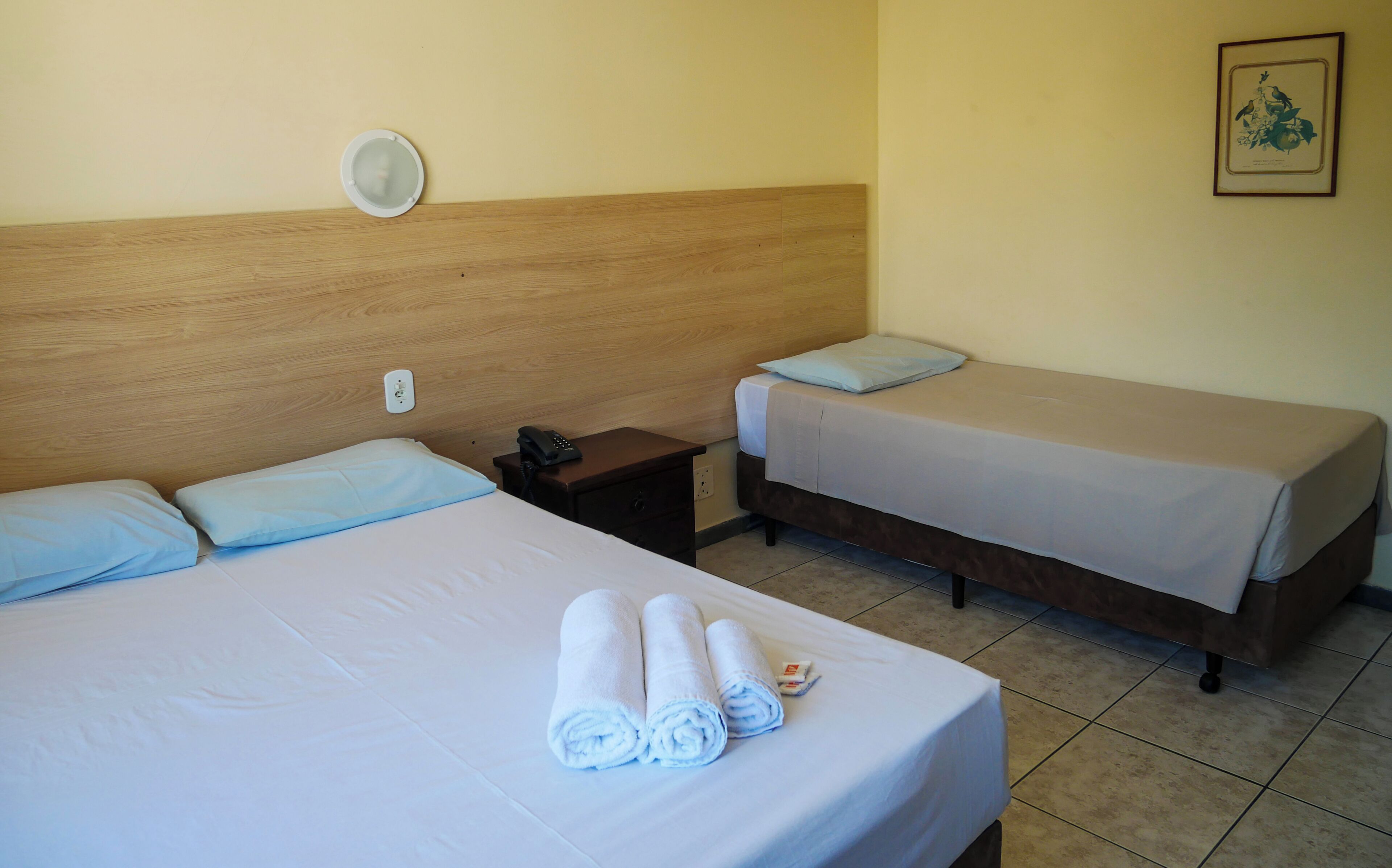 A hotel room with two beds, a bedside table with a phone, a wall socket, and a framed picture.