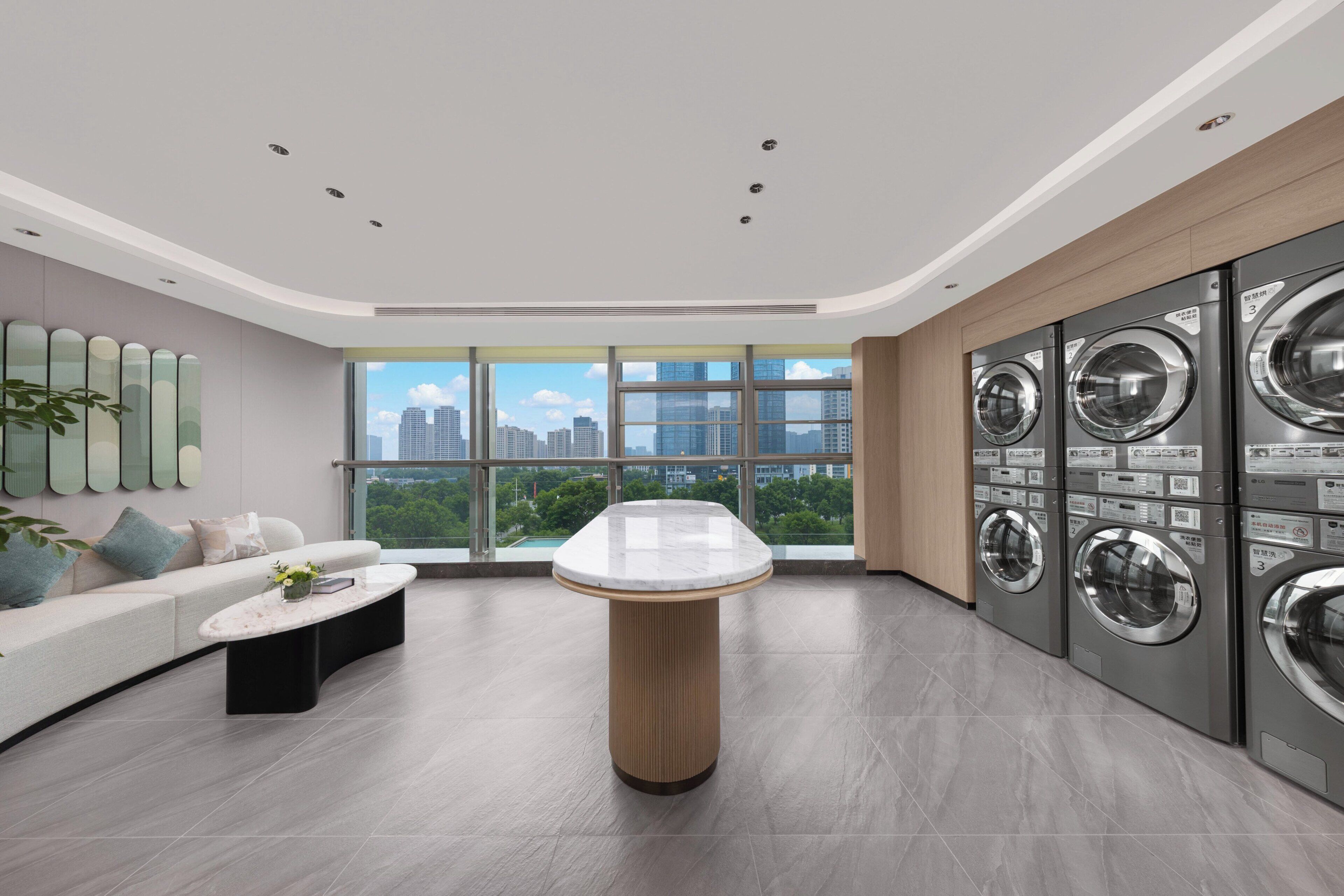 A modern interior with a curved coffee table, a sofa with cushions, a washing machine unit, and a large window offering a city view.