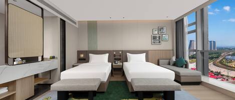 Quarto de hotel moderno com duas camas, uma janela grande com vista para a cidade, uma escrivaninha com cadeira e um pequeno espaço para sentar.